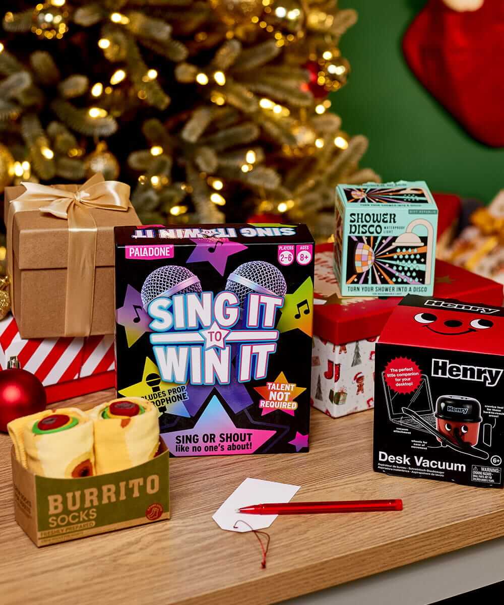 Selection of novelty Christmas gifts, including a singing game, desk vacuum, shower disco gadget and burrito socks, displayed on a table in front of a decorated Christmas tree. Selection of novelty Christmas gifts, including a singing game, desk vacuum, shower disco gadget and burrito socks, displayed on a table in front of a decorated Christmas tree.